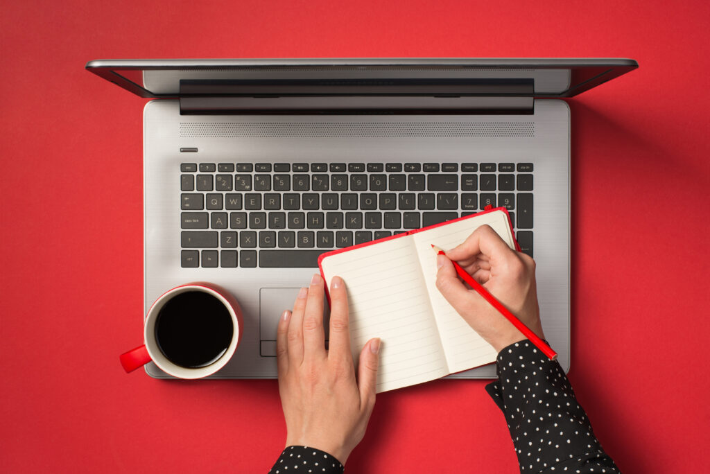 first-person-top-view-photo-female-hands-writing-with-red-pencil-red-notepad-cup-coffee-laptop-isolated-red-background